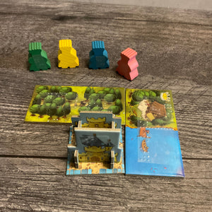 The meeples with braille on them. in front of them is the starting castle, and two domino tiles with forest and water. All are labeled in braille