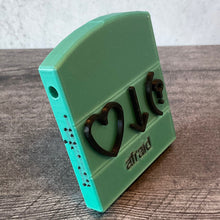 Load image into Gallery viewer, Afraid bliss symbols. This is an example but all words are this same design. Front and side view. Side has braille in black and a hole for a string. Base is color is teal. Front says afraid in print and has the 3 symbols that Volk uses for this word, heart, down arrow and parenthesis with question mark.

