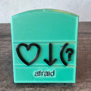 Afraid bliss symbol. This is an example but all words are this same design. Front view. Base is color is teal. Front says afraid in print and has the 3 symbols that Volk uses for this word, heart, down arrow and parenthesis with question mark.
