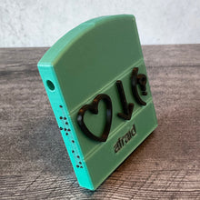 Load image into Gallery viewer, Afraid bliss symbols. This is an example but all words are this same design. Front and side view. Side has braille in black and a hole for a string. Base is color is teal. Front says afraid in print and has the 3 symbols that Volk uses for this word, heart, down arrow and parenthesis with question mark.
