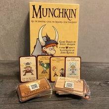 Load image into Gallery viewer, Munchkin box with 4 cards showing face up and 2 piles of cards in front of them. the face up cards have braille stickers. The piles of cards have QR codes on the back.
